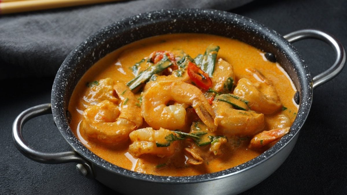Eine Pfanne mit dem Curry auf einem grauen Tisch. Im dunklen Hintergrund sind unscharf Holz-Stäbchen zu erkennen. Alles von schräg oben fotografiert.