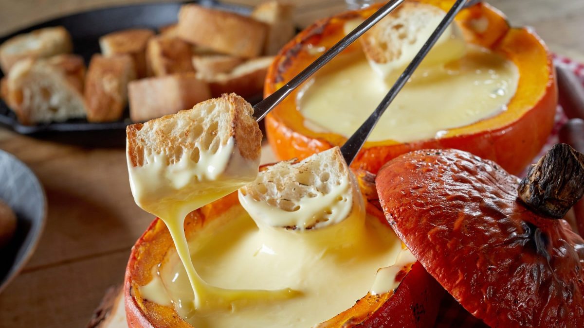 Zwei Kürbisse mit Käse-Fondue auf einem Holztisch seitlich fotografiert. Daneben noch eine gusseiserne Pfanne mit Brotstückchen.