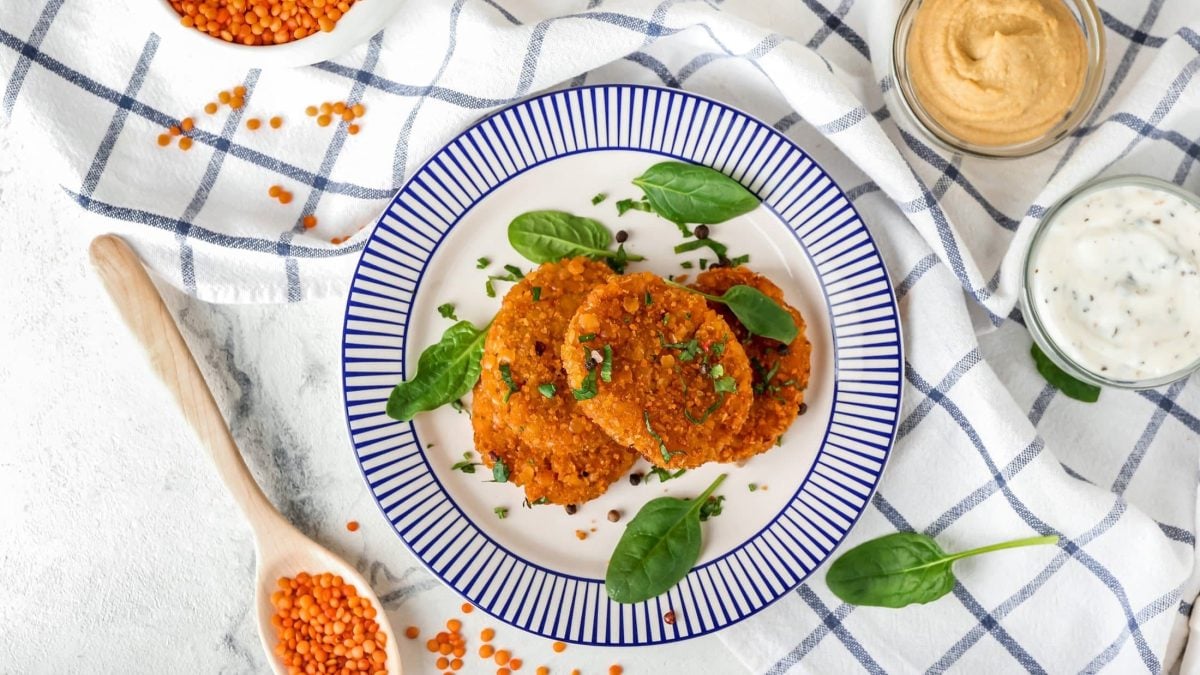 Draufsicht: Auf einem hellen Untergrund steht ein weißer Teller mit blauem Rand. Auf dem liegen vegane Linsen-Frikadellen. Drum herum sind zwei Schalen mit Dips und Spinatblätter.