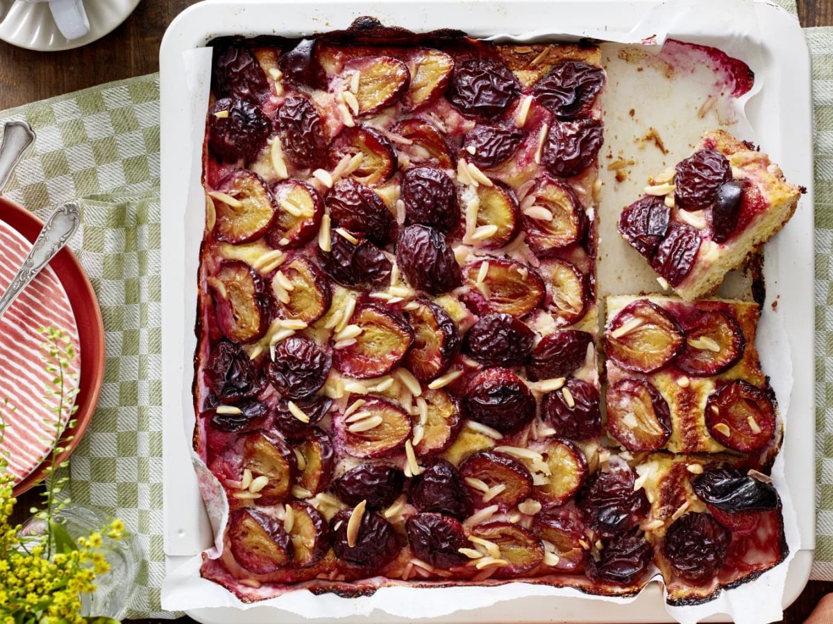 Ein Marzipan-Pflaumenkuchen in einem weißen Blech auf einem grün-weiß-kariertem Geschirrtuch auf einem hölzernen Hintergrund. Links davon Teller mit Dessertgabeln, rechts eine kleine Schüssel mit Mandelstiften.