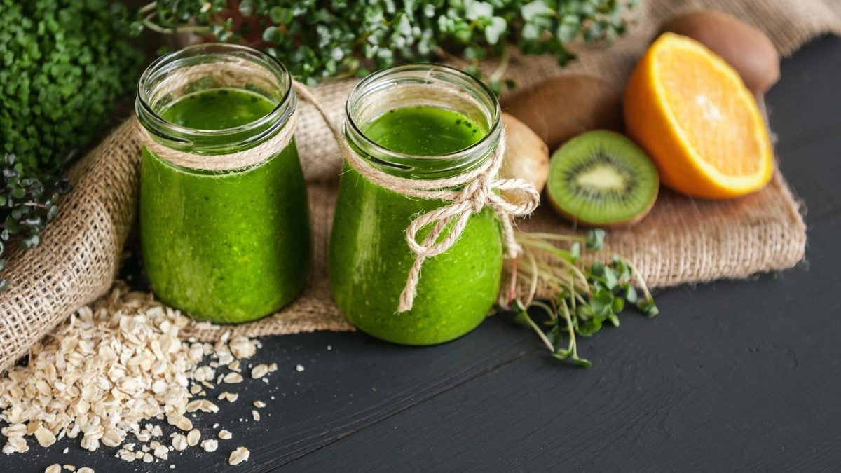 Zwei Gläser Microgreen-Smoothies auf einem dunklem Untergrund. Drum herum liegen Sprossen, Orangen und Kiwis.