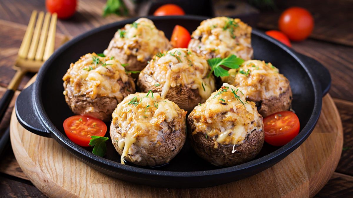 Draufsicht, schräg: eine gusseiserne Pfanne mit Ofen-Champignons mit Knofi-Hack-Füllung auf einem Holzbrett. Daneben liegt eine Gabel.
