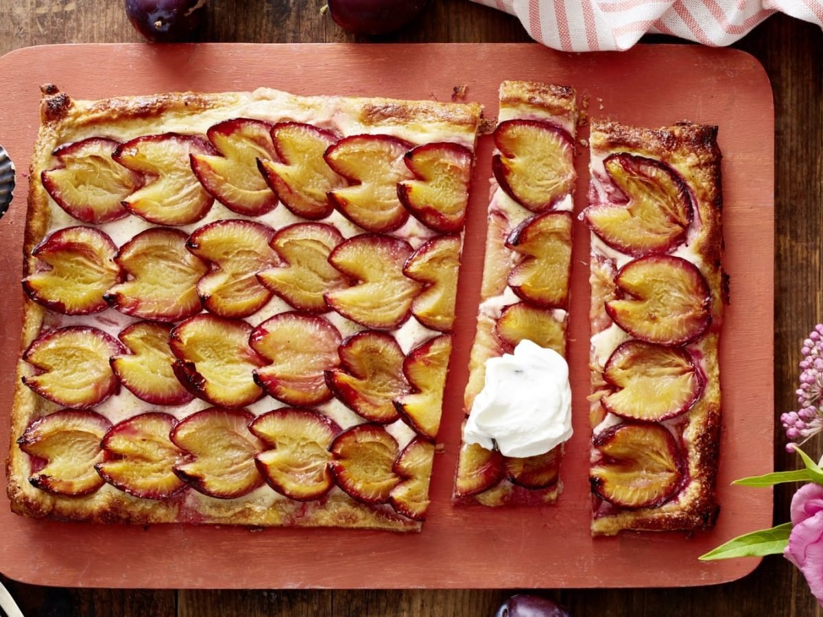 Eine Pflaumen-Blätterteig-Tarte auf einem roten Schneidebrett. Links davon eine Schüssel mit Schlagsahne und zwei Dessertgabeln. Rechts ein Geschirrtuch und Blumen. Oben auf dem Bild Pflaumen. Der Hintergrund ist hölzern.