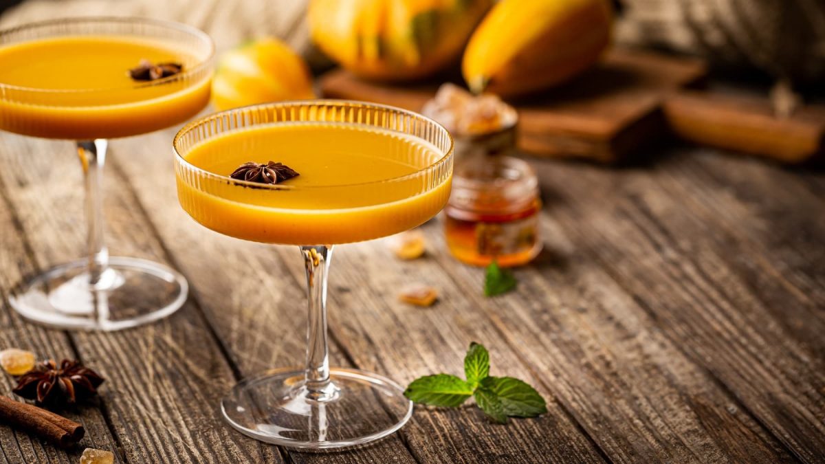 Zwei Gläser mit dem Pumpkin Passion auf einem Holztisch mit herbstlicher Deko (Kürbis und Gewürze). Alles von leicht schräg oben fotografiert.