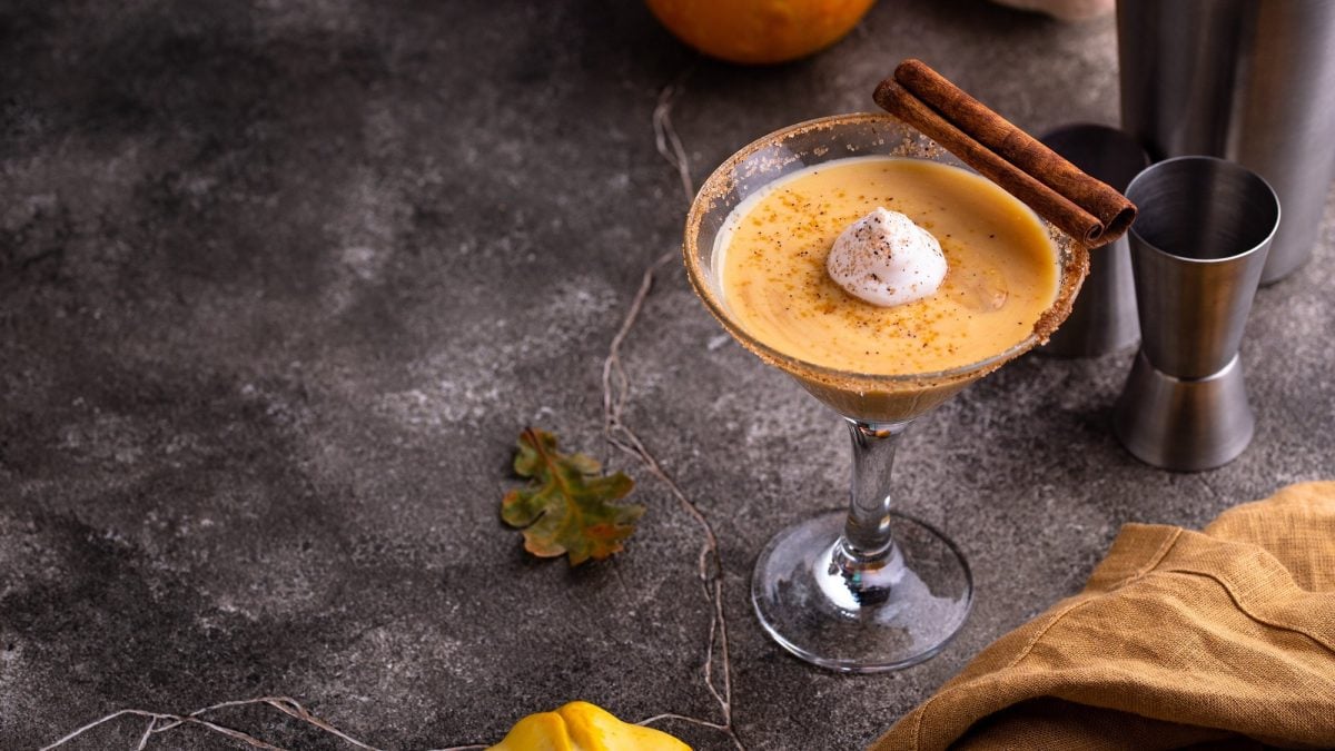 Ein Martiniglas mit Pumpkin Spice Likör und einem Klecks Sahne, garniert mit Zimtstangen, auf dunklem Untergrund, daneben ein Kürbis.