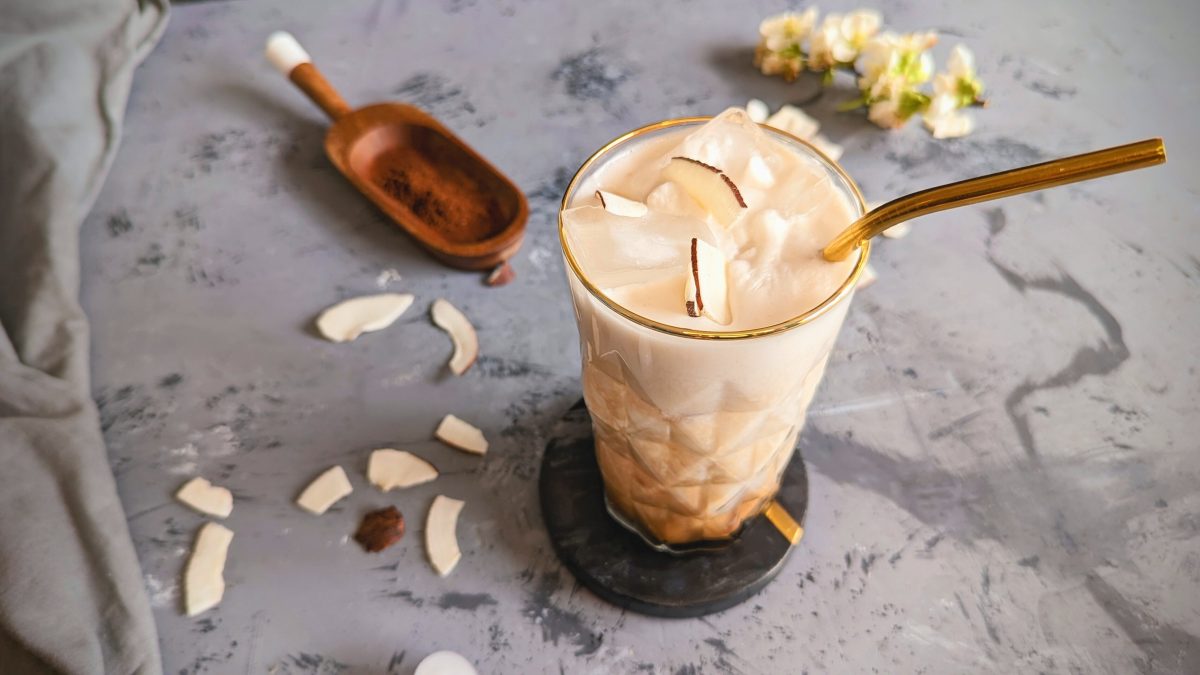 Ein Glas mit dem Bounty-Latte auf einem grauen Steintisch mit Kokosflocken und weißen Blüten. Alles von schräg oben fotografiert.