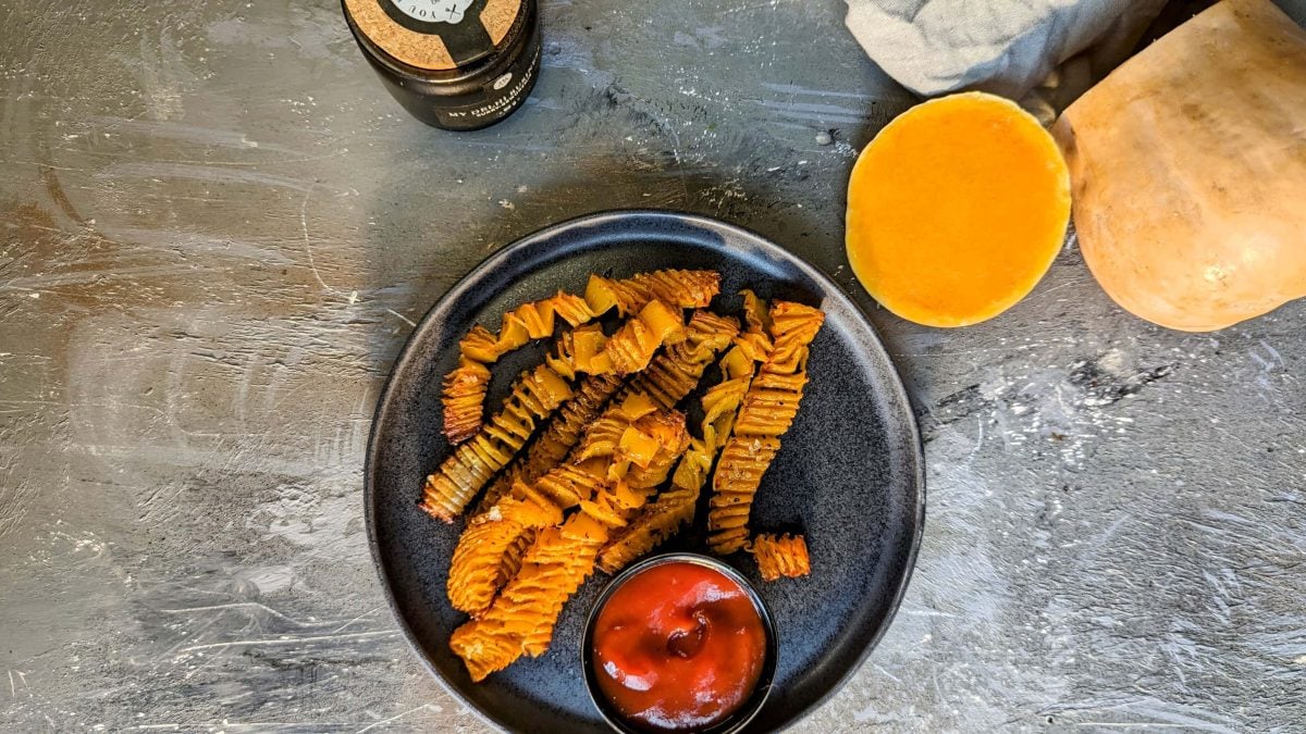 Ein schwarzer Teller mit den Kürbis-Akkordeon-Pommes und Ketchup auf einem grauen Steintisch. Alles direkt von oben fotografiert.