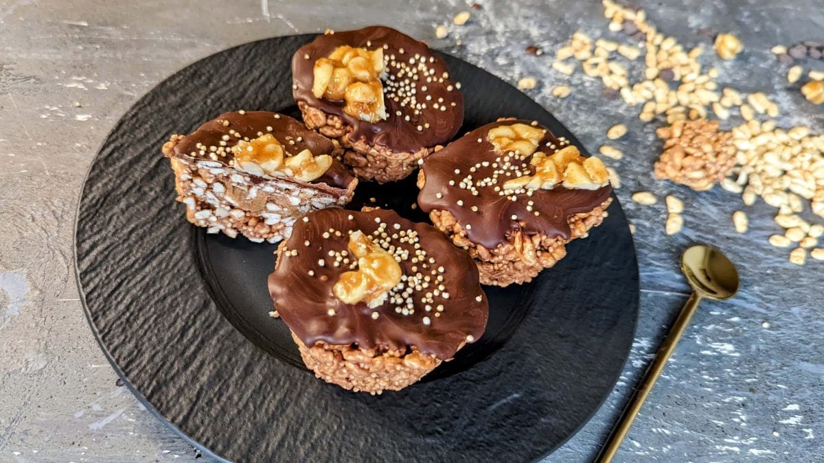 Vier Peanutbutter Rice-Cups auf einem schwarzen Teller und grauem Untergrund. Daneben gepuffter Reis und ein Löffel mit Erdnussbutter.