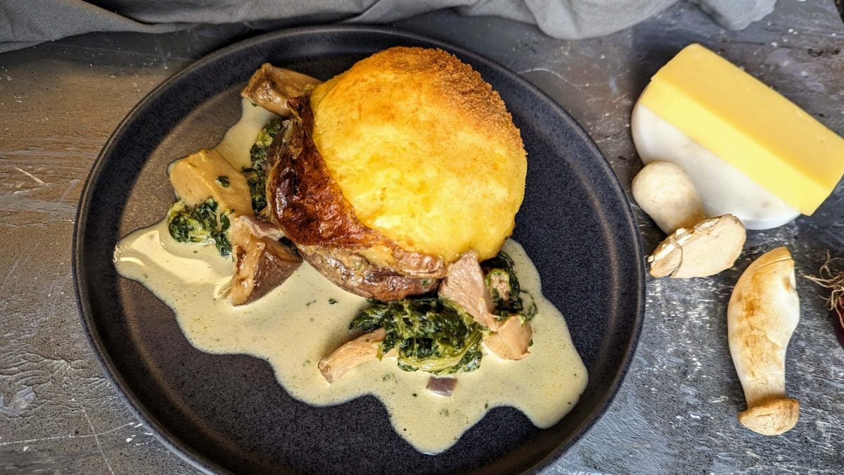Ein schwarzer Teller mit dem Käse-Pilz-Knödel auf einem grauen Tisch von oben fotografiert.