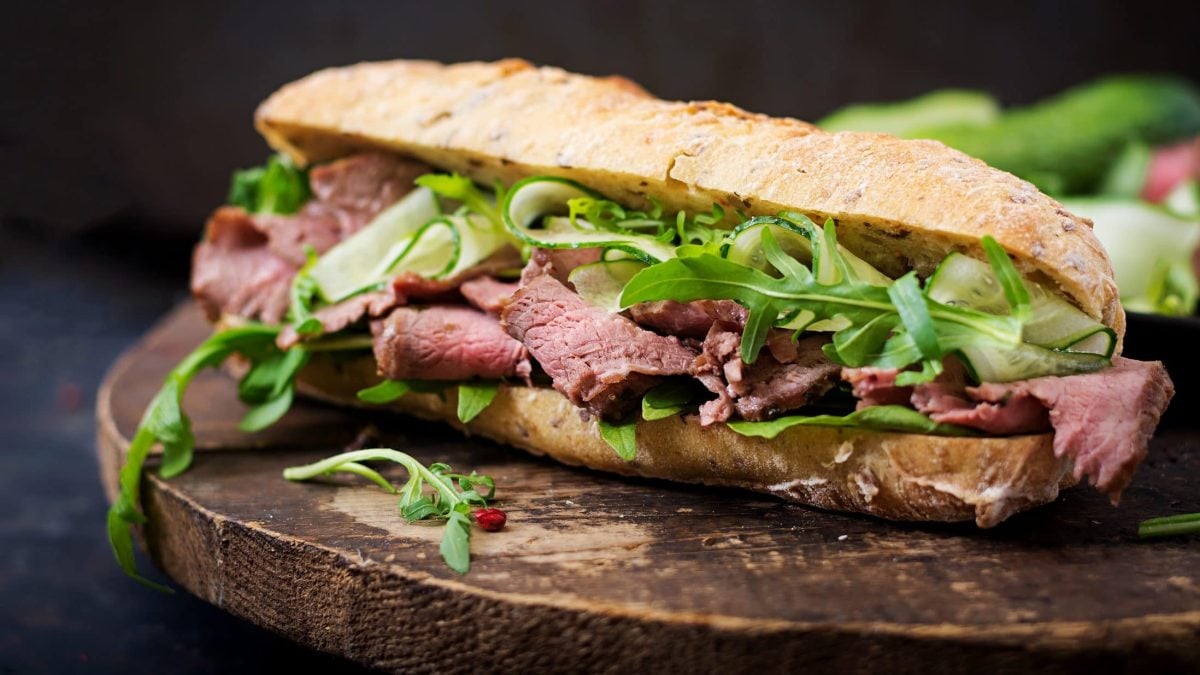 Frontal: ein Roastbeef-Gurken-Sandwich vor dunklem Hintergrund auf einem runden Holztisch.
