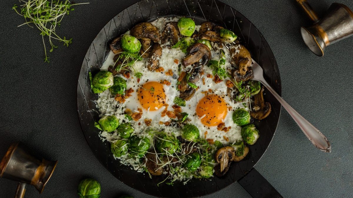 Draufsicht: eine Rosenkohl-Champignon-Pfanne mit zwei Spiegeleiern auf einem dunklen Untergrund. Daneben liegen Sprossen und ein Besteck.