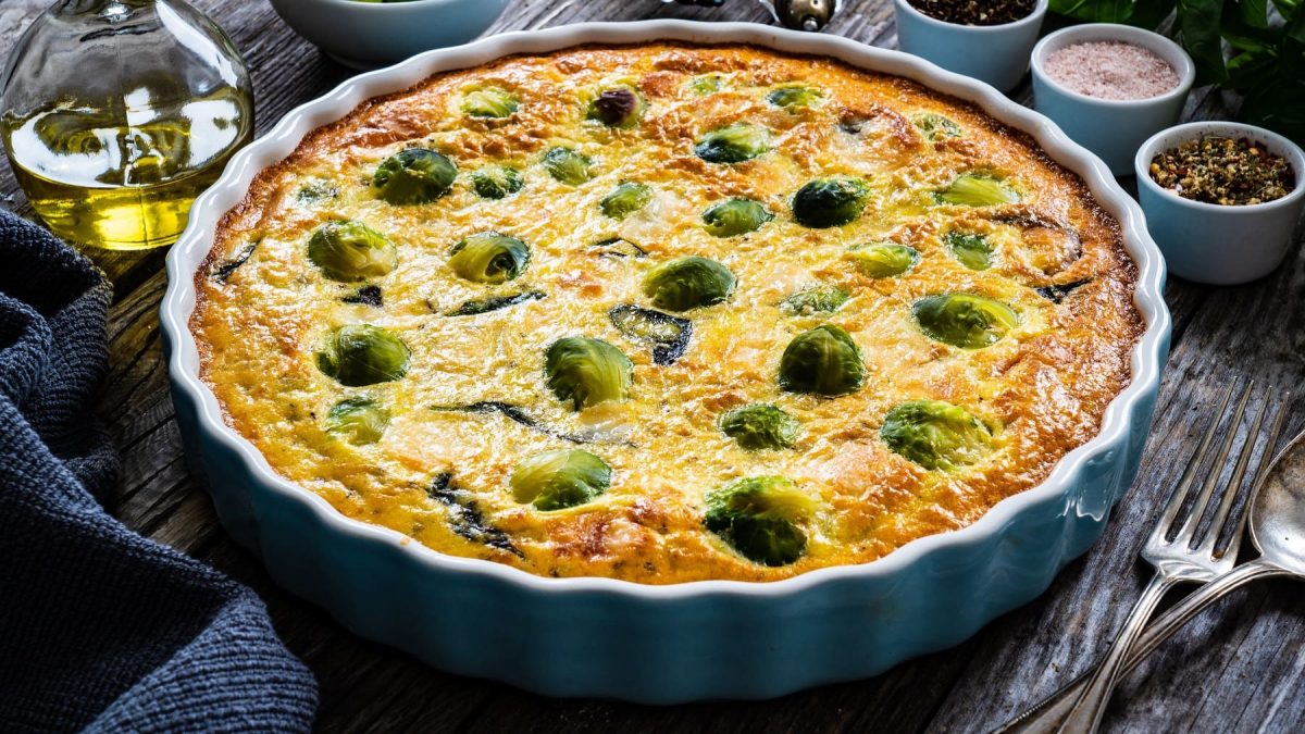 Eine blaue Auflaufform mit der Rosenkohl-Quiche auf einem dunklen Holztisch von schräg oben fotografiert.