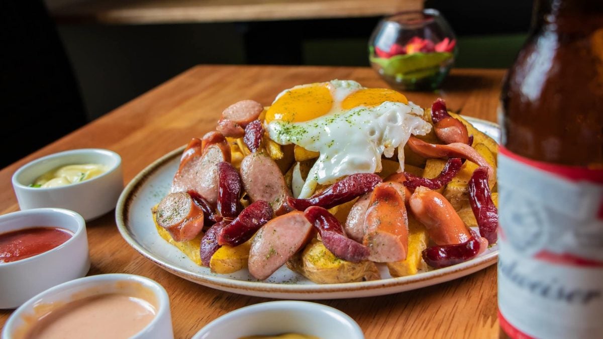 Draufsicht schräg oben: Ein Teller mit Bratkartoffeln, gebratenen Wiener Würstchen und einem Spiegel obenauf steht auf einem Tisch. Drum herum sind viele Schalen mit unterschiedlichen Saucen. / Salchipapa