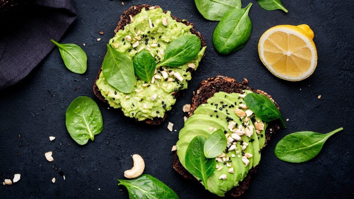 Draufsicht: Schwarzer Untergrund, zwei Sandwich mit Avocado-Cashew-Creme, Spinatblätter, halbierte Zitrone.
