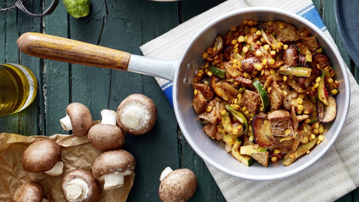 Eine Schnitzel-Gemüse-Pfanne mit Linsen auf einem Geschirrtuch auf einem dunkelblauen Hintergrund. Links unten Champignons und eine Flasche Öl.