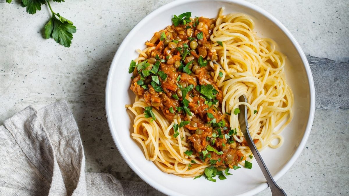 Ein weißer Teller mit Spaghetti und Linsen-Bolognese auf einem grauen Steintisch direkt von Oben fotografiert. Links vom Bild außerdem, Petersilie und ein graues Tuch. Rechts im Bild eine silberne Gabel, die in den Teller ragt.