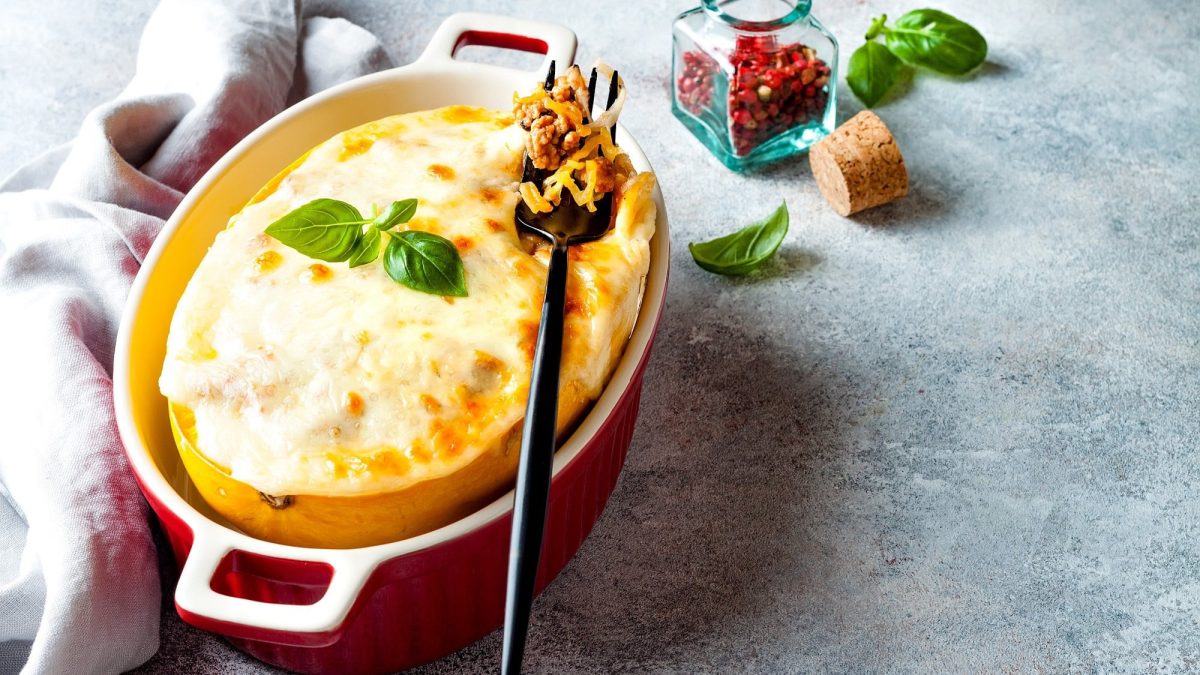 Ein halber Spaghettikürbis Elsässer Art in einer roten Auflaufform, mit Käse überbacken und Basilikum garniert, darauf eine Gabel, drumherum etwas frischer Basilikum und ein Glas mit rotem Pfeffer.