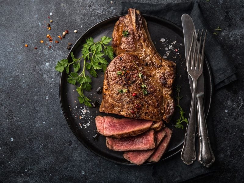 Ein schwarzer Teller mit einem perfekt gegartem Steak auf dunklem Untergrund mit Salz, Gewürzen und Kräutern direkt von oben fotografiert.