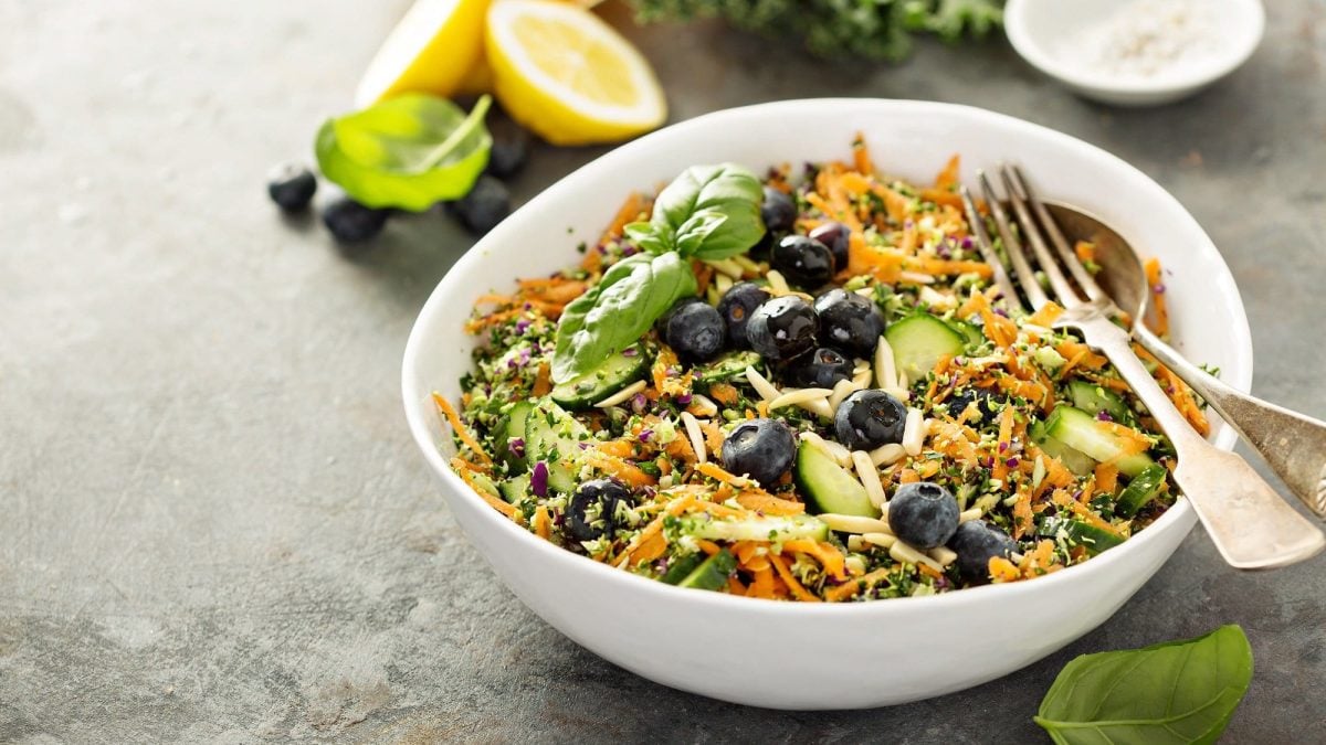 Eine Schüssel Superfood-Salat mit geraspelter Möhre, zerkleinertem Brokkoli, Heidelbeeren, Gurke, darin Besteck, daneben eine aufgeschnittene Zitrone.