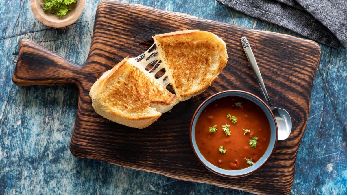 Ein Toast mit veganem Mozzarella gefüllt auf einem hölzernen Schneidebrett auf einem dunkelgrauen Hintergrund. Auch noch auf dem Brett eine kleine Tomatensuppe und ein Löffel. Links oben ein Schüsselchen mit Petersilie.