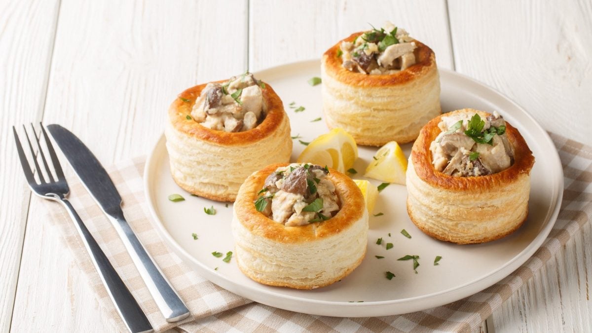 4 Vol-au-vent mit Pilzen auf einem weißen Teller, dazwischen Zitronenspalten, daneben Messer und Gabel sowie eine Serviette.