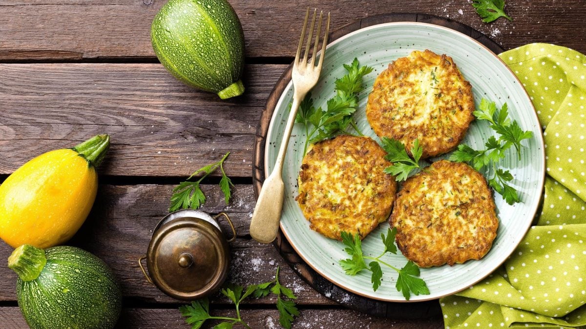 Draufsicht: Auf einem Holztisch steht ein Teller mit Zucchini-Puffer mit Hüttenkäse. Drum herum liegen Zucchini und ein gelbes Tuch.