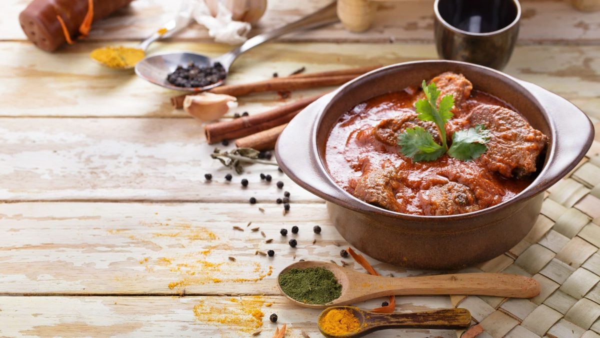 Draufsicht schräg: Ein Keramiktopf asiatisches Lammcurry, dass auch Rogan Josh genannt wird, steht auf einem Untergrund aus Holz. Drum herum liegen Gewürzen.