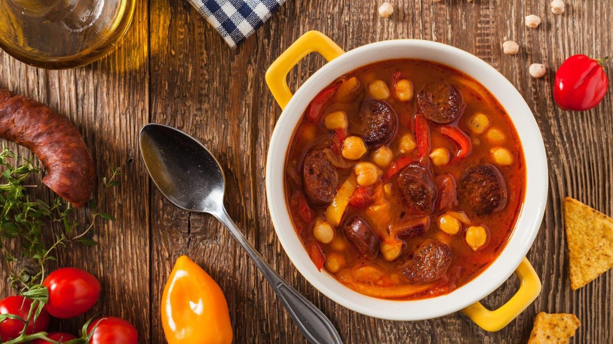 Draufsicht: ein gelber Topf mit einer Chorizo-Suppe mit Kichererbsen steht auf einem Untergrund aus Holz. Drum herum liegen rote Tomaten und gelbe Paprika.