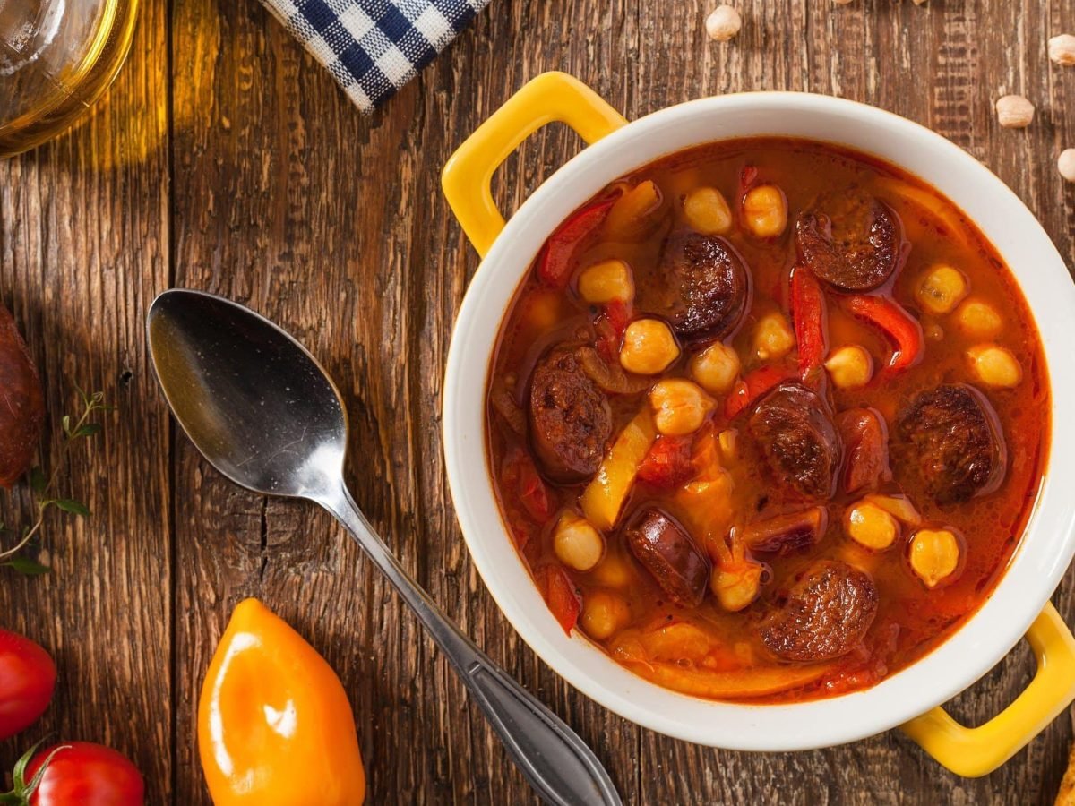 Draufsicht: ein gelber Topf mit einer Chorizo-Suppe mit Kichererbsen steht auf einem Untergrund aus Holz. Drum herum liegen rote Tomaten und gelbe Paprika.