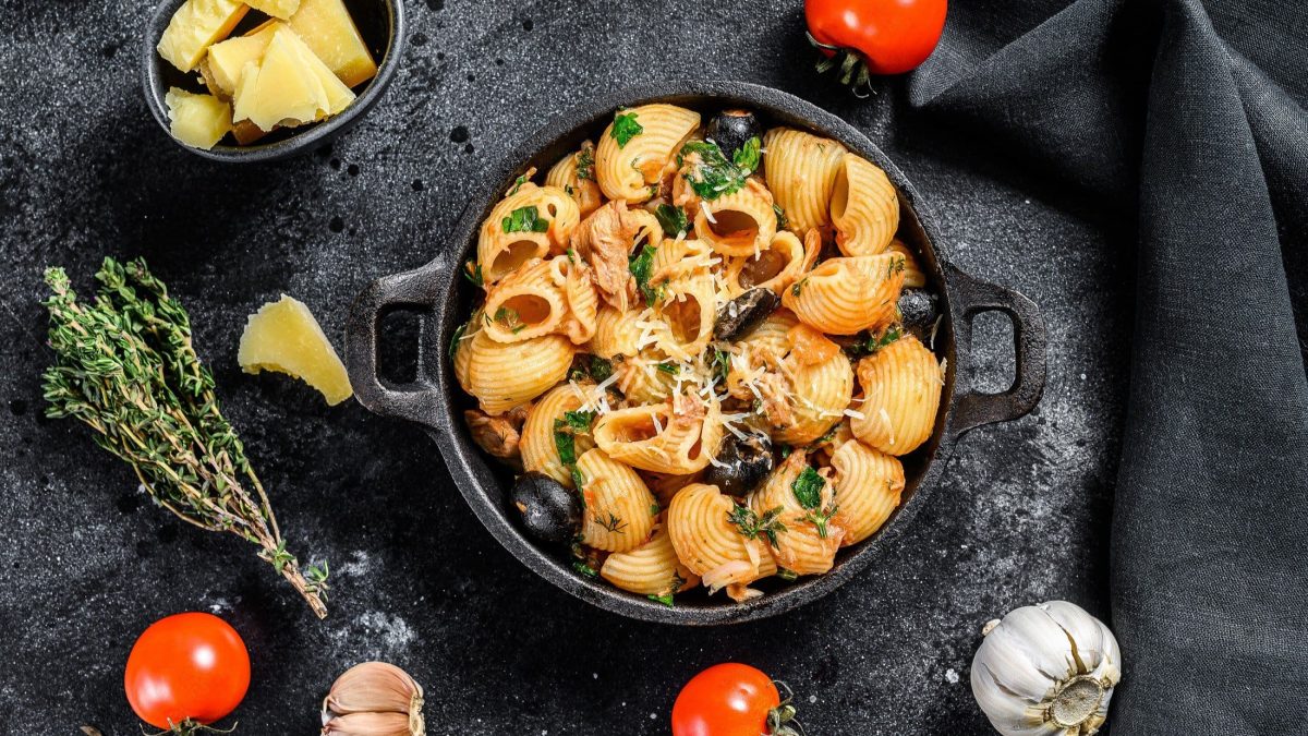 Draufsicht: Eine dunkle Pfanne mit Conchiglie Rigate mit Thunfisch steht auf einem dunklem Untergrund. Drum liegen Tomaten, Knoblauch und Gewürze.