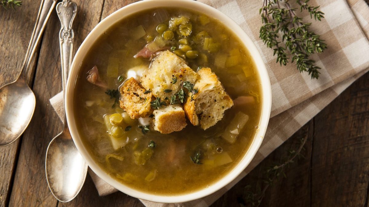Eine weiße Suppenschale mit der Erbsen-Lauch-Suppe auf einem Holztisch mit frischem Thymian und einer weiß-braun-karierten Tischdecke und zwei Löffeln. Alles von oben fotografiert.