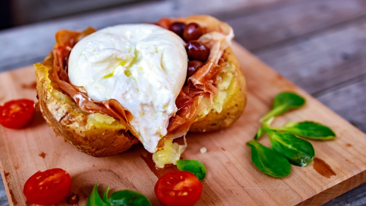 Eine gebackene und halbierte Kartoffel mit Burrata, Schinken und Oliven gefüllt auf einem Holzbrett mit Kirschtomaten und Feldsalat.