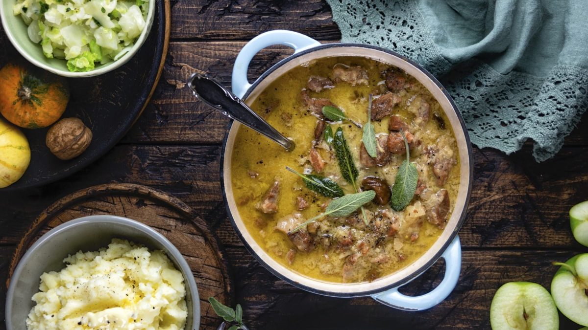 Geschmortes Schweinefleisch mit Äpfeln in einem Schmortopf. Rechts oben ein dunkelgrünes Geschirrtuch, links eine Schüssel mit Kartoffelpüree, links oben eine Schüssel mit Wirsinggemüse. Rechts unten auf dem Bild drei Apfelhälften.