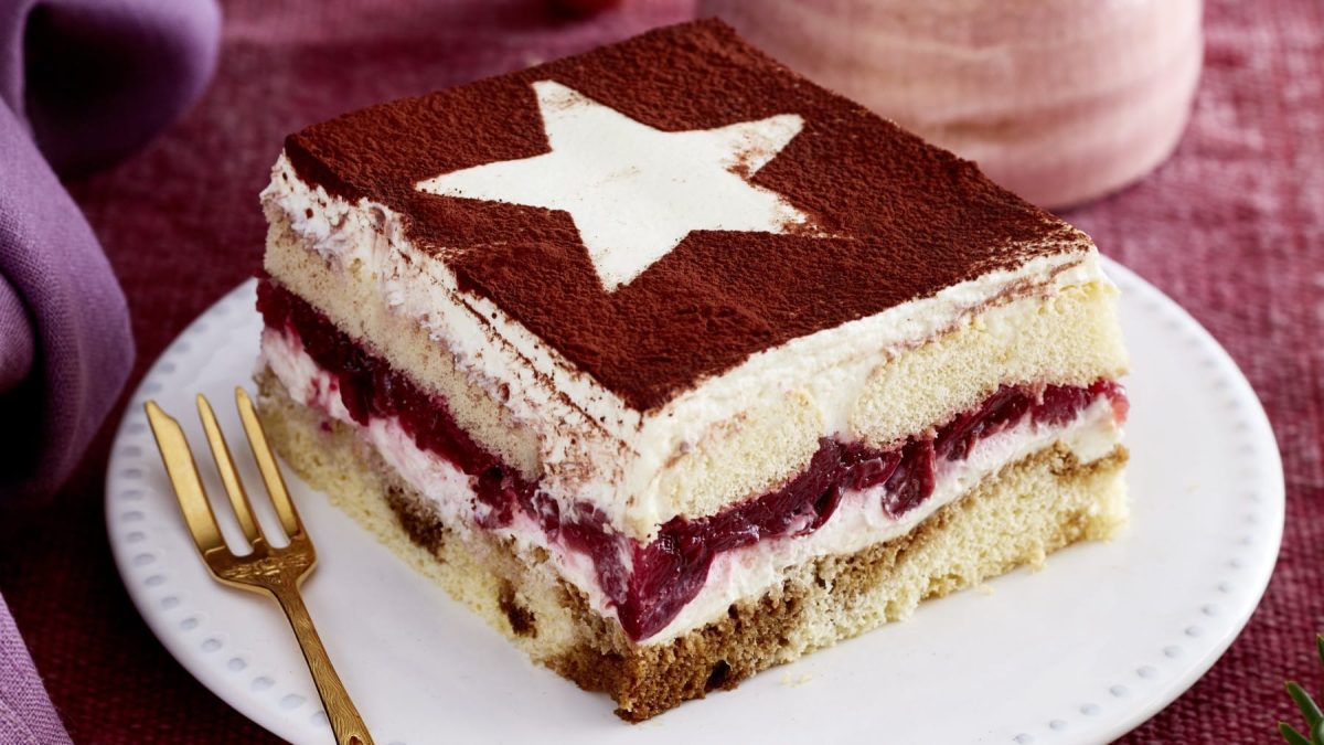 Ein Stück Glühwein-Tiramisu auf einem weißen Teller mit einer goldenen Gabel. Der Hintergrund ist rot.