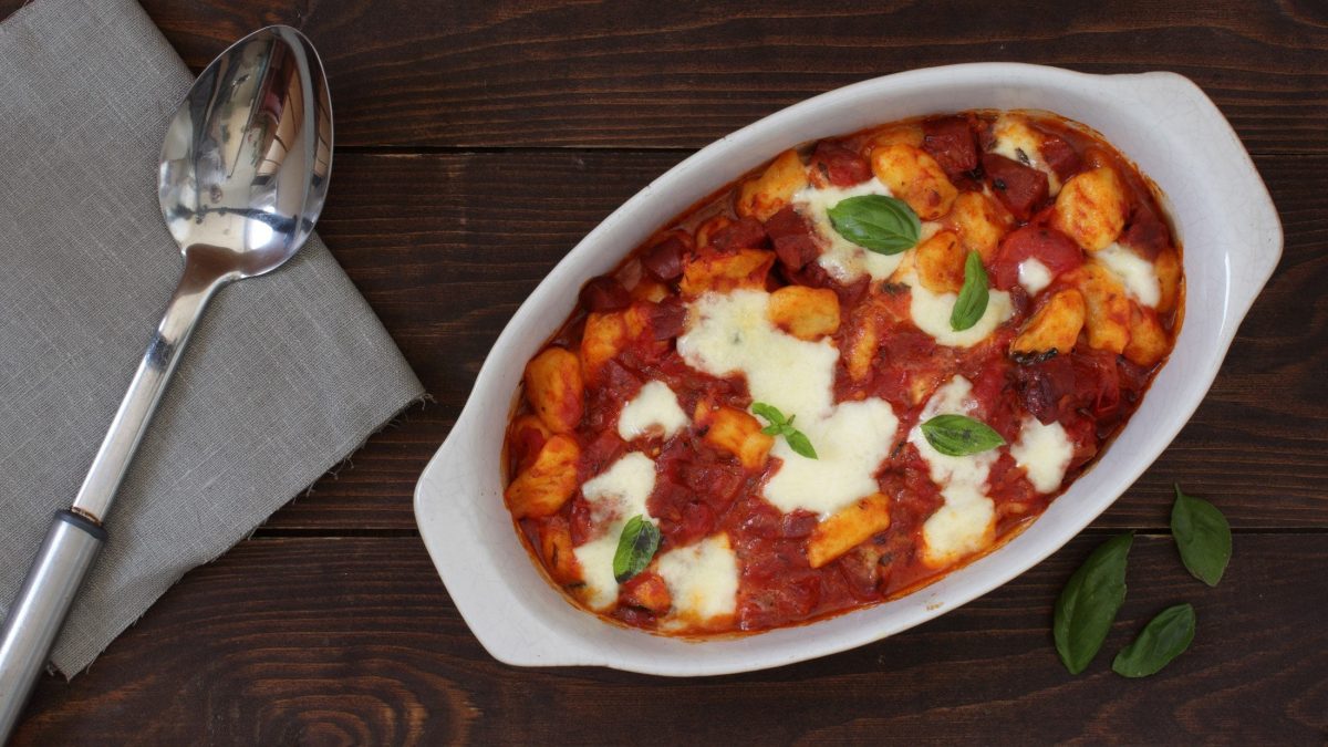 Draufsicht: ein Gnocchi-Chorizo-Auflauf auf einem dunklen Untergrund. Daneben liegen ein Löffel und eine Stoffserviette.
