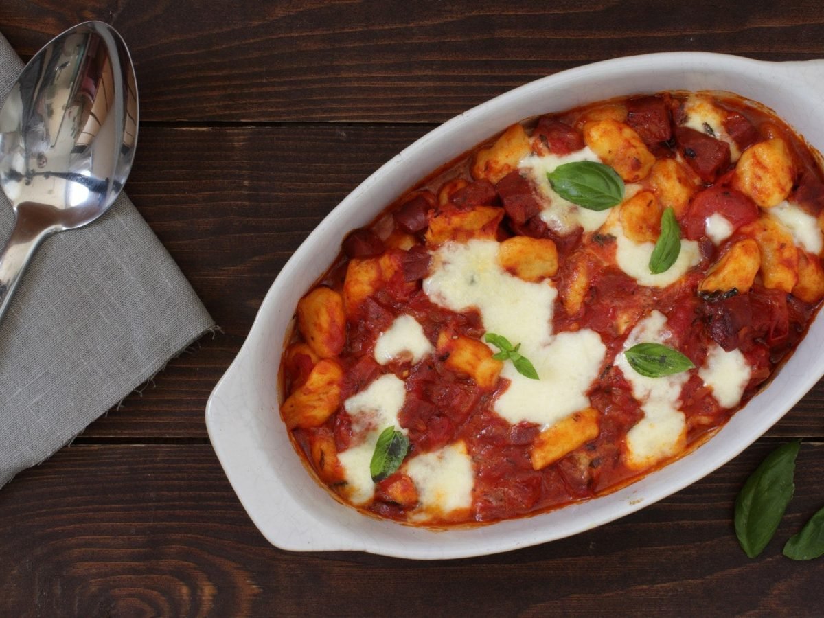 Draufsicht: ein Gnocchi-Chorizo-Auflauf auf einem dunklen Untergrund. Daneben liegen ein Löffel und eine Stoffserviette.