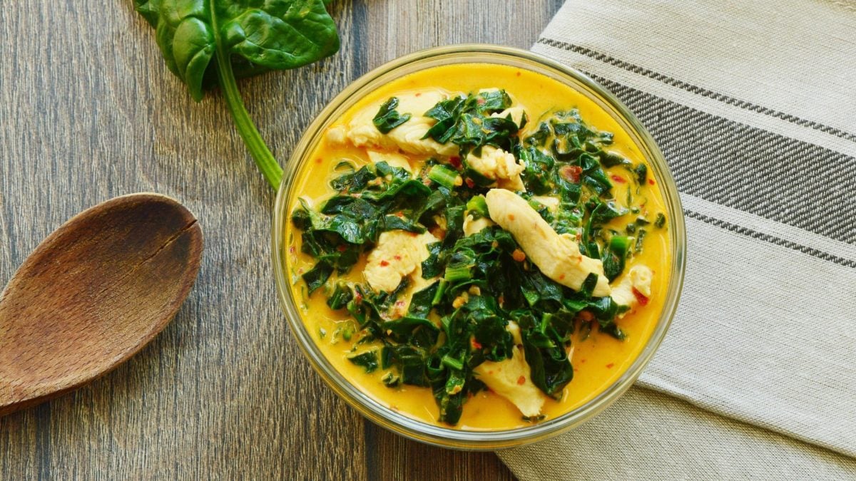 Draufsicht: eine Schüssel mit einem Hähnchen-Curry mit Spinat steht auf einem Tuche, das auf Holz lieht. Daneben sind ein Spinatblatt und ein Löffel aus Holz.