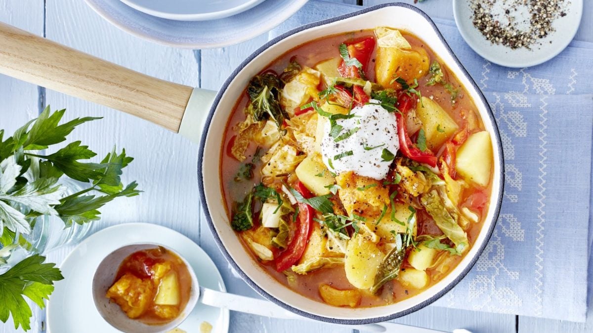 Draufsicht: Kartoffel-Wirsing-Gulasch im Topf auf hellblauem Untergrund, drum herum Geschirr und Zutaten.