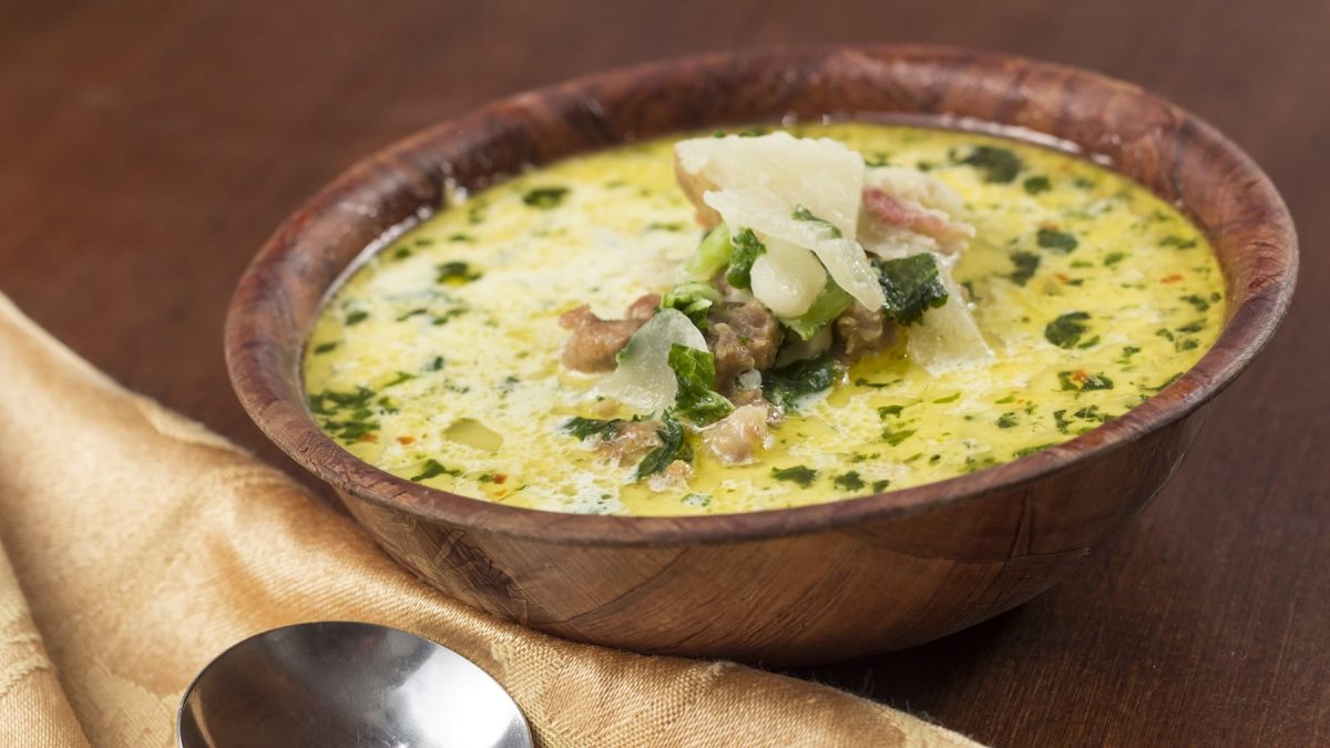 Eine braune Holzschale mit einer Kohlsuppe mit Käse und Hackfleisch auf einem hellbraunen Tisch. Alles von der Seite fotografiert.