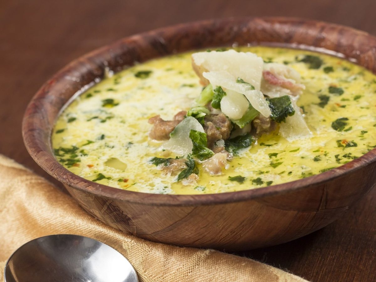 Eine braune Holzschale mit einer Kohlsuppe mit Käse und Hackfleisch auf einem hellbraunen Tisch. Alles von der Seite fotografiert.
