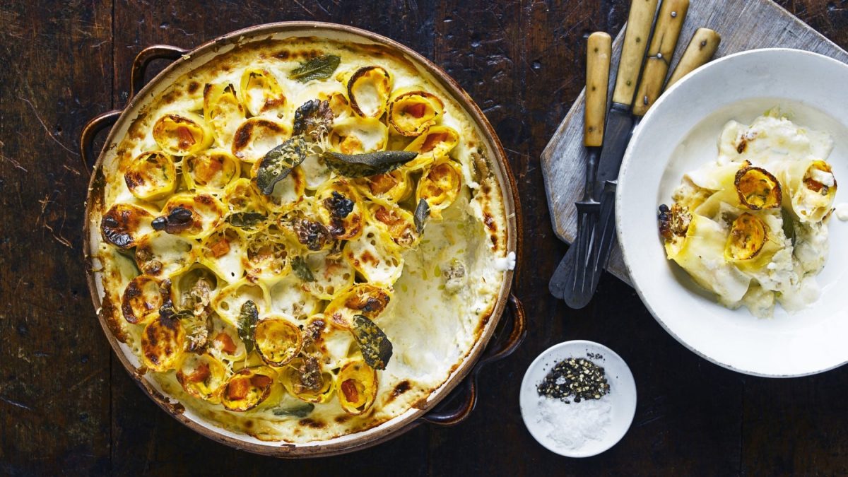 Kürbis-Ricotta-Cannelloni mit Salbei in einer Auflaufform auf einem hölzernen Hintergrund. Rechts davon ein Teller mit einer Portion Cannelloni, Besteck und ein kleines Schüsselchen mit Salz und Pfeffer.