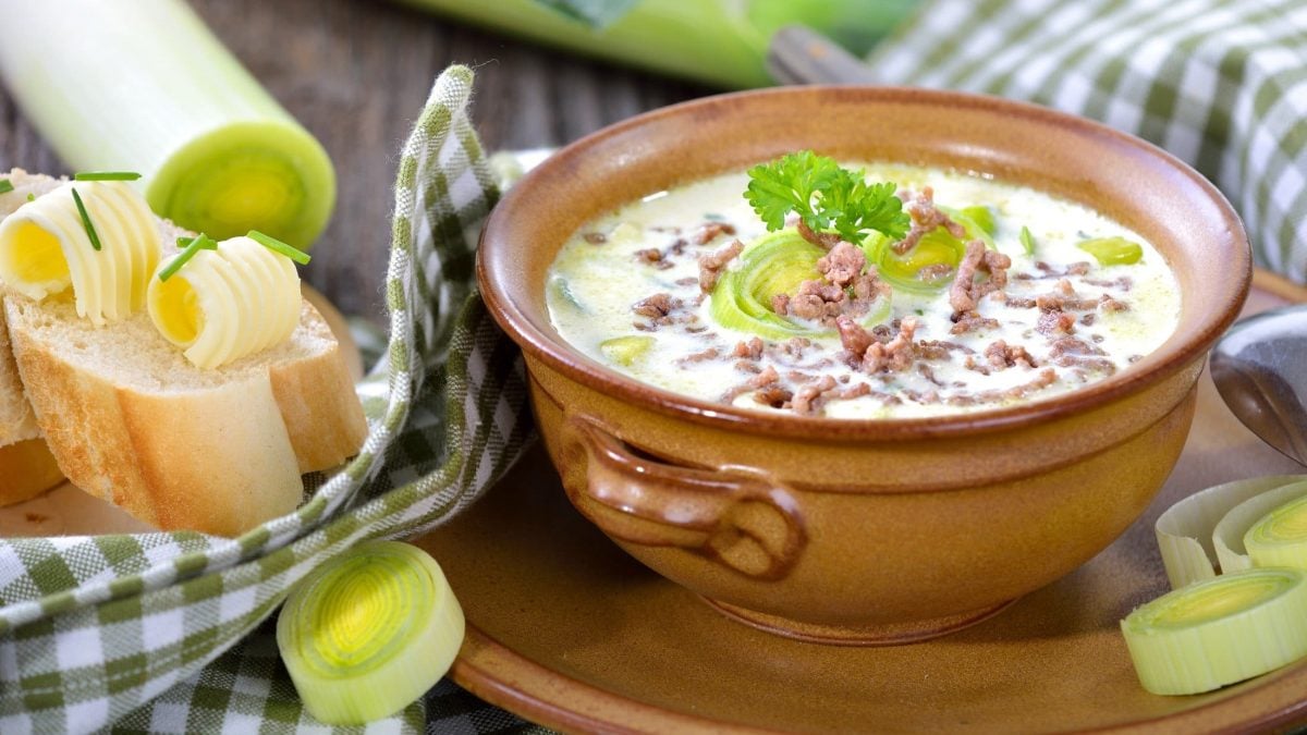 Draufsicht, schräg: Eine Ton-Schüssel mit einer Lauchsuppe mit Hackfleisch. Drum herum liegen Lauchstangen, Lauchringe sowie ein eine Scheibe Weißbrot mit Butter.