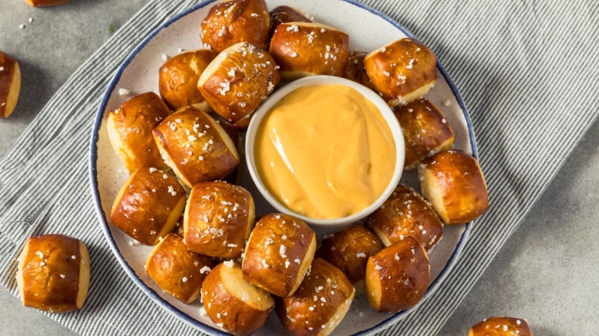 Brezel-Bites auf einem weißen Teller mit blauen Rand, in der Mitte ein Schüsselchen mit Käsesauce. Der Teller steht auf einem grau-weiß-gestreiften Geschirrtuch, der Hintergrund ist grau.