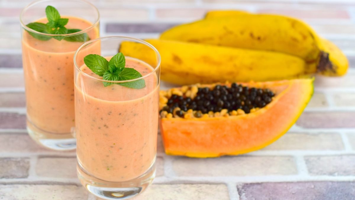 Zwei kleine Gläser mit dem Papaya Shake auf einem Ziegelsteinboden. Daneben ein Stück frische Papaya und 3 Bananen. Alles seitlich und nah fotografiert.