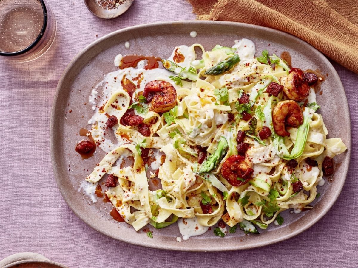 Pasta in Mandel-Lemon-Cream mit Garnelen und Chorizo auf rosa Teller mit gleichem Untergrund, drum herum Gläser und Küchentuch in ockergelb.