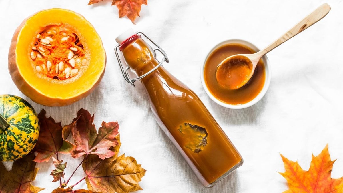 Eine kleine Glasflasche mit dem Sirup auf weißem Untergrund. Daneben Herbstlaub und verschiedene Kürbisse. Alles direkt von oben fotografiert.