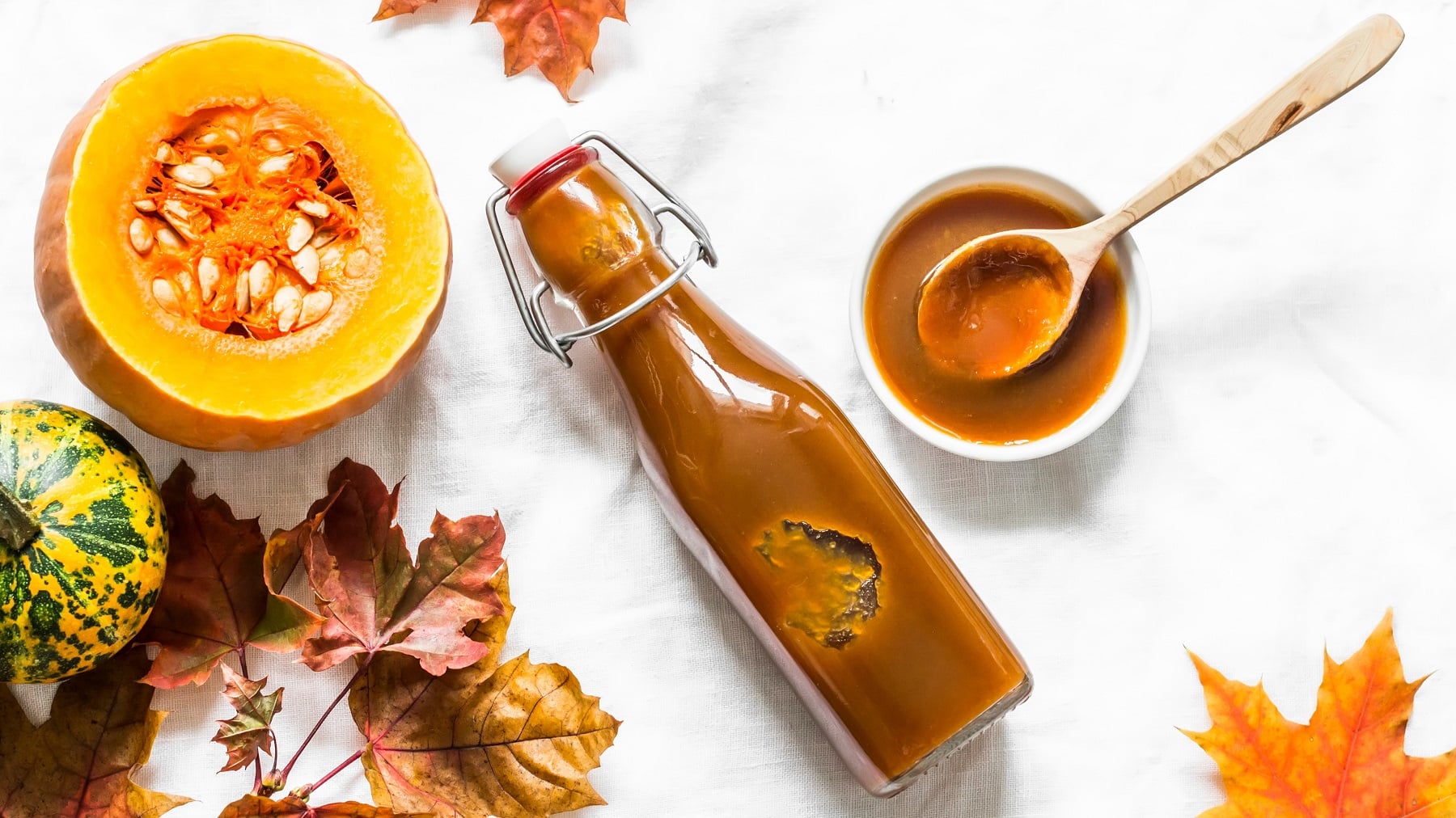 Eine kleine Glasflasche mit dem Sirup auf weißem Untergrund. Daneben Herbstlaub und verschiedene Kürbisse. Alles direkt von oben fotografiert.