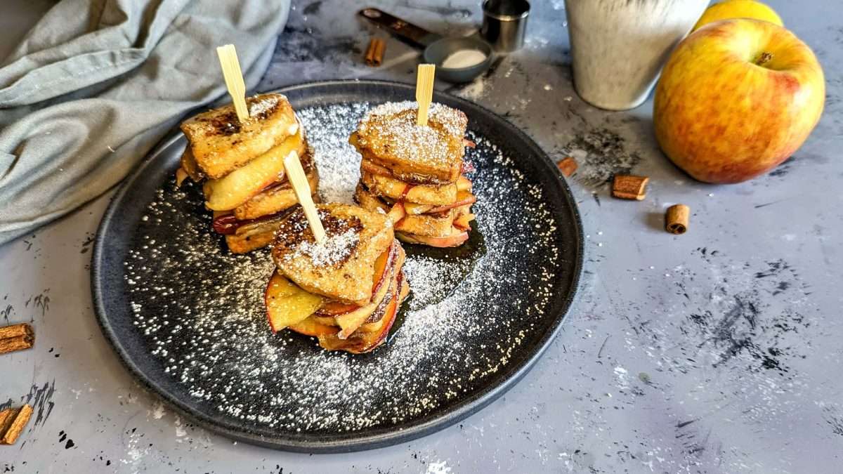 Ein schwarzer Teller mit den Mini-French-Toasts mit Aofel auf einem grauen Tisch mit winterlicher Stimmung. Alles von leicht schräg oben fotografiert.
