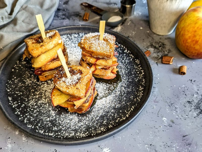 Ein schwarzer Teller mit den Mini-French-Toasts mit Aofel auf einem grauen Tisch mit winterlicher Stimmung. Alles von leicht schräg oben fotografiert.