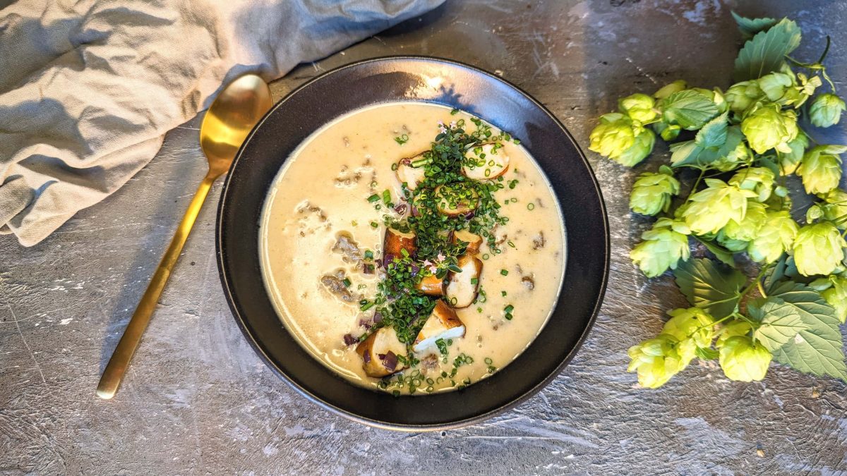 Eine schwarze Schüssel mit der Suppe auf einem grauen Untergrund. Daneben frischer Hopfen und ein goldener Löffel. Alles direkt von oben fotografiert.